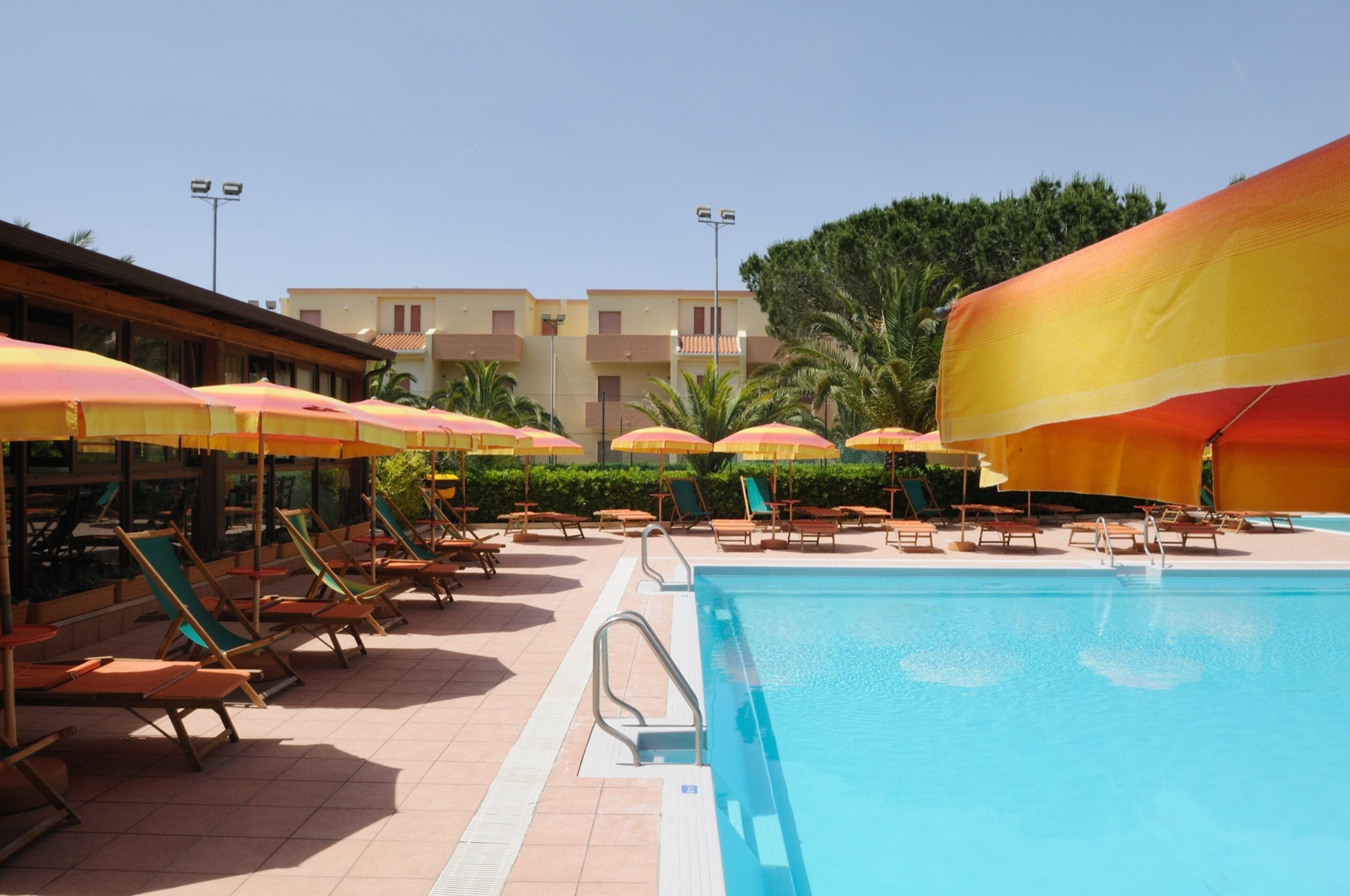 A poolside area with sun loungers and umbrellas, a clear blue pool, and a building with balconies in the background.