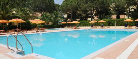 Una piscina amb gandules i ombrel·les, envoltada d'arbres i un solàrium enrajolat.