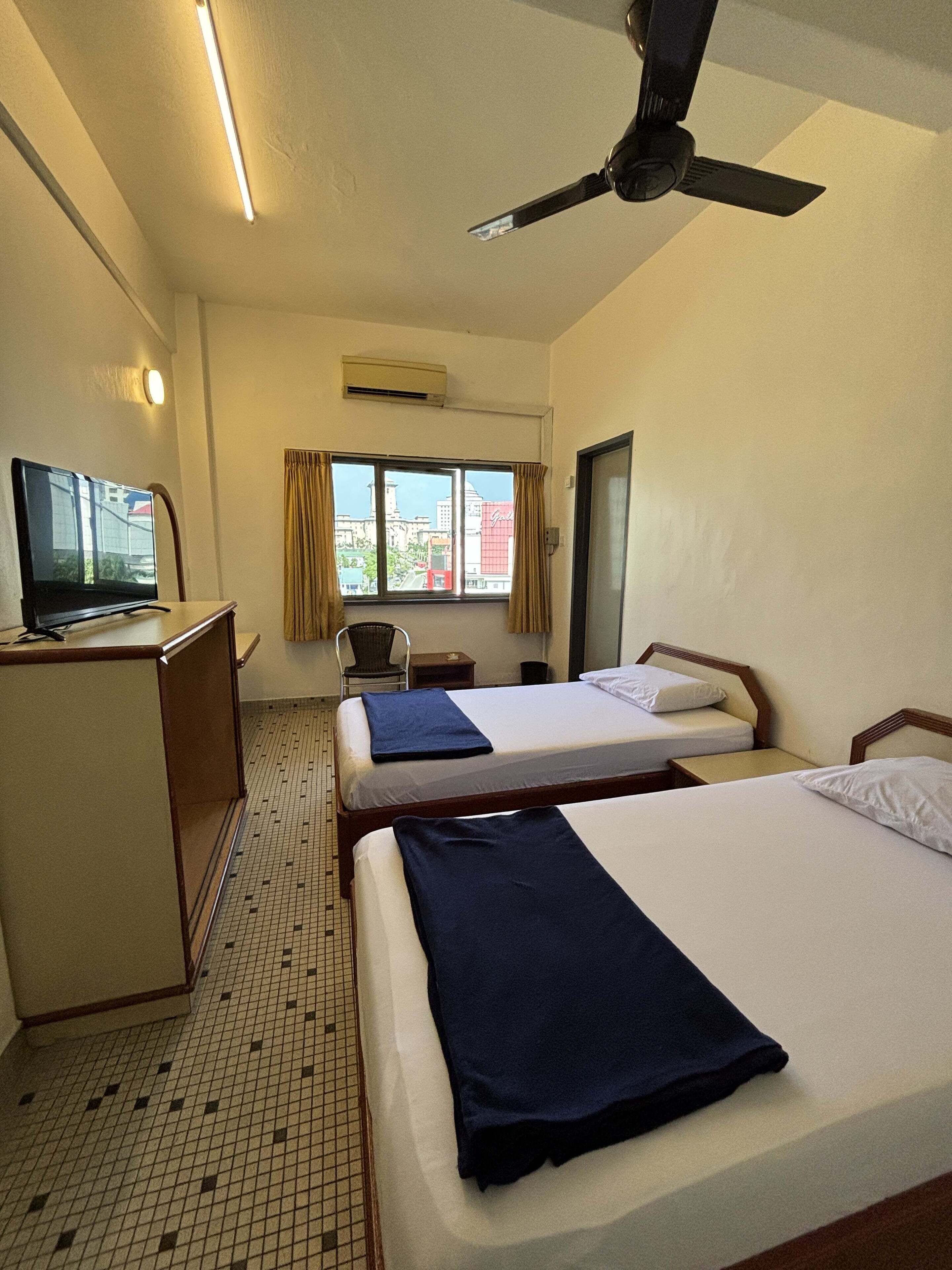 A hotel room with two beds, a desk with a television, a ceiling fan, and a window with curtains.