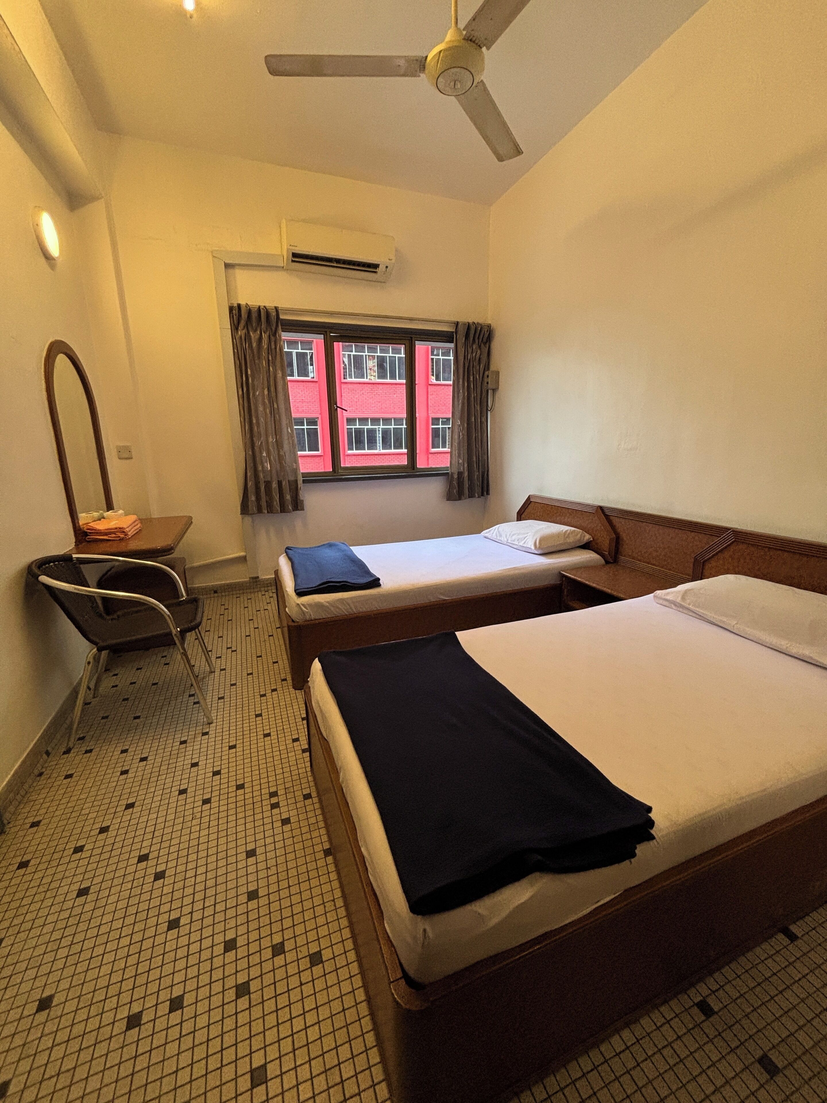 A hotel room with two beds, a chair, a ceiling fan, an air conditioning unit, and a window with curtains.