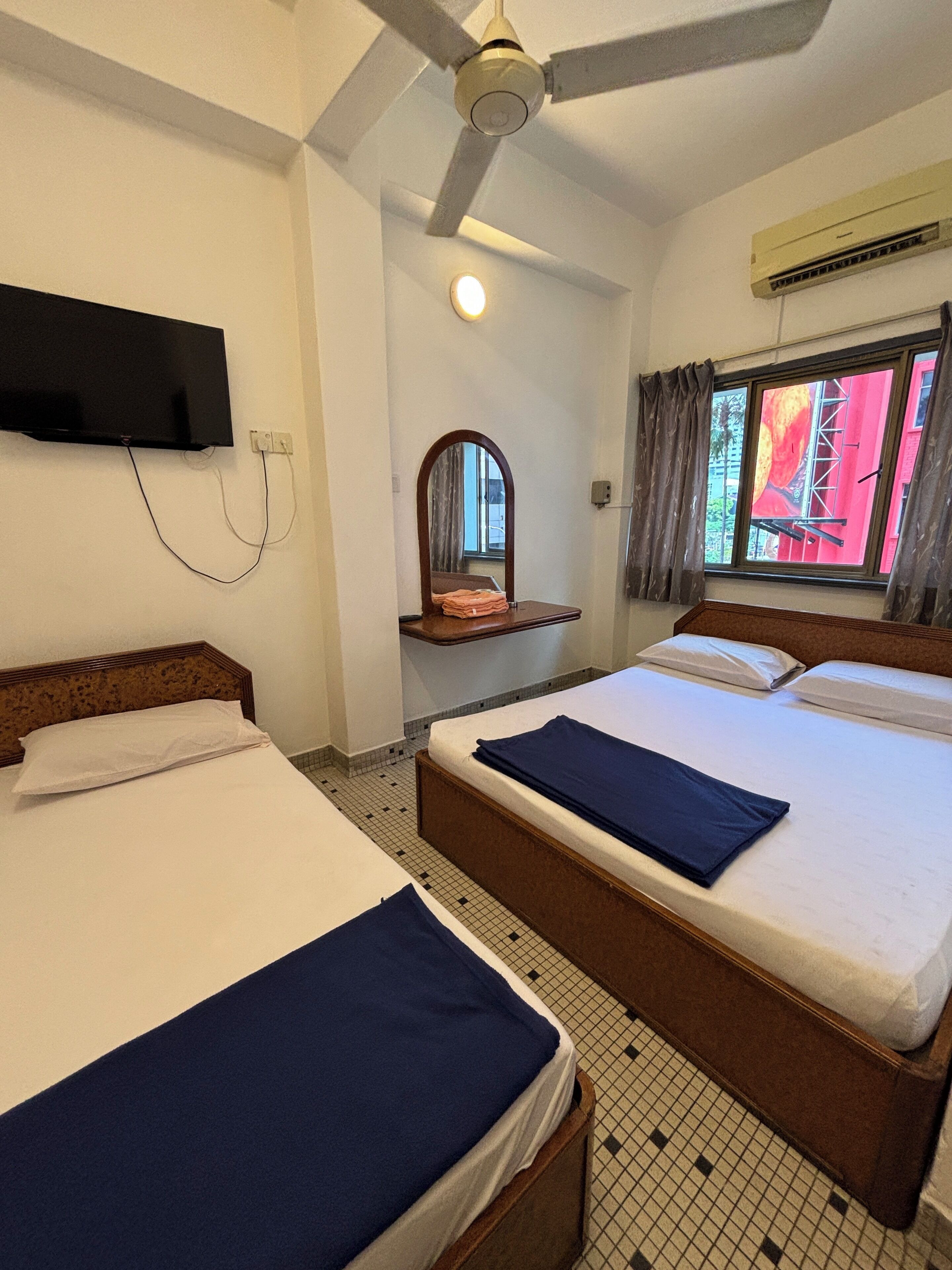 A hotel room with two beds, a ceiling fan, a mounted TV, a window with curtains, and an air conditioning unit.