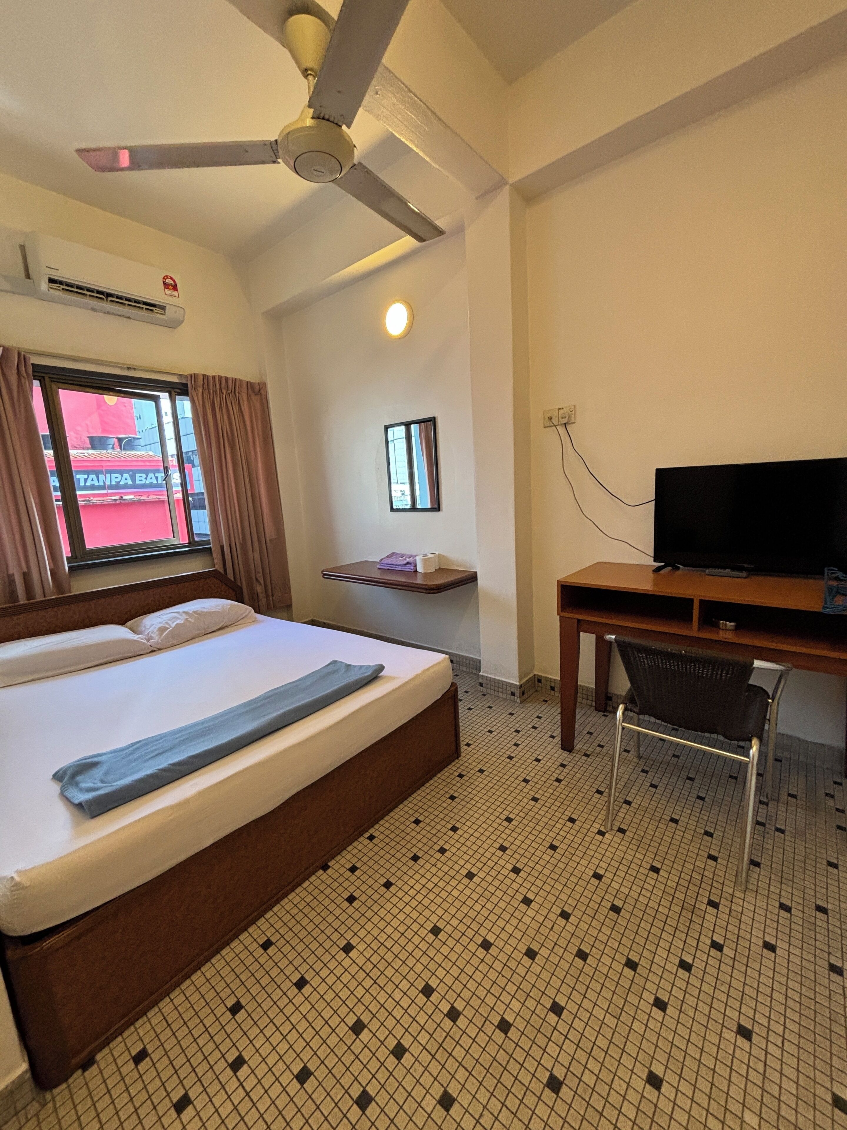 A hotel room with a bed, a desk, a chair, a ceiling fan, a window with curtains, a small shelf, and a television.