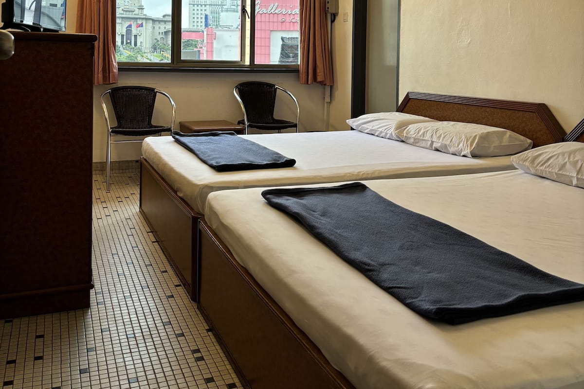 A hotel room with two beds, a desk, a chair, a window with curtains, and a ceiling fan.