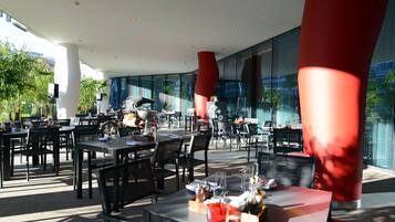 Un área de comedor al aire libre con mesas y sillas, ventanas amplias y elementos arquitectónicos modernos.