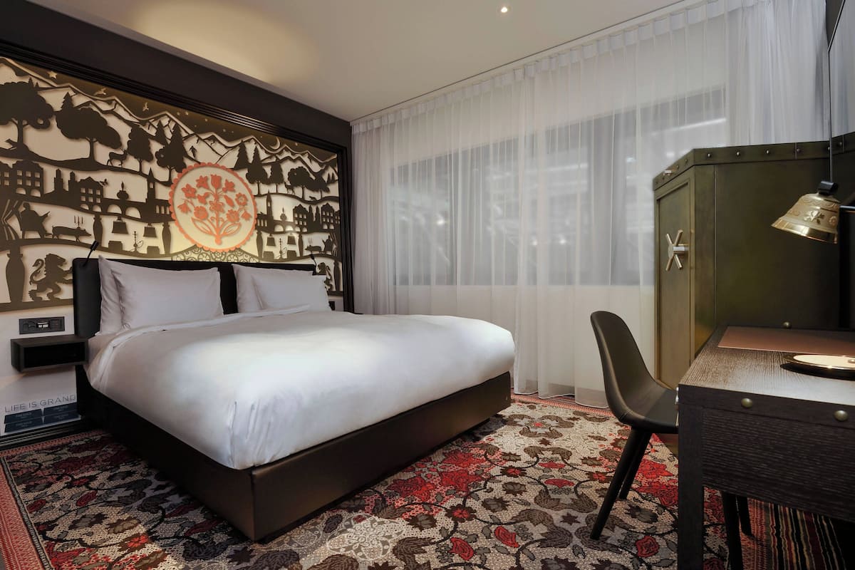 A hotel room with a large bed, a desk, and a chair. There is a decorative wall with a tree and a flower design.