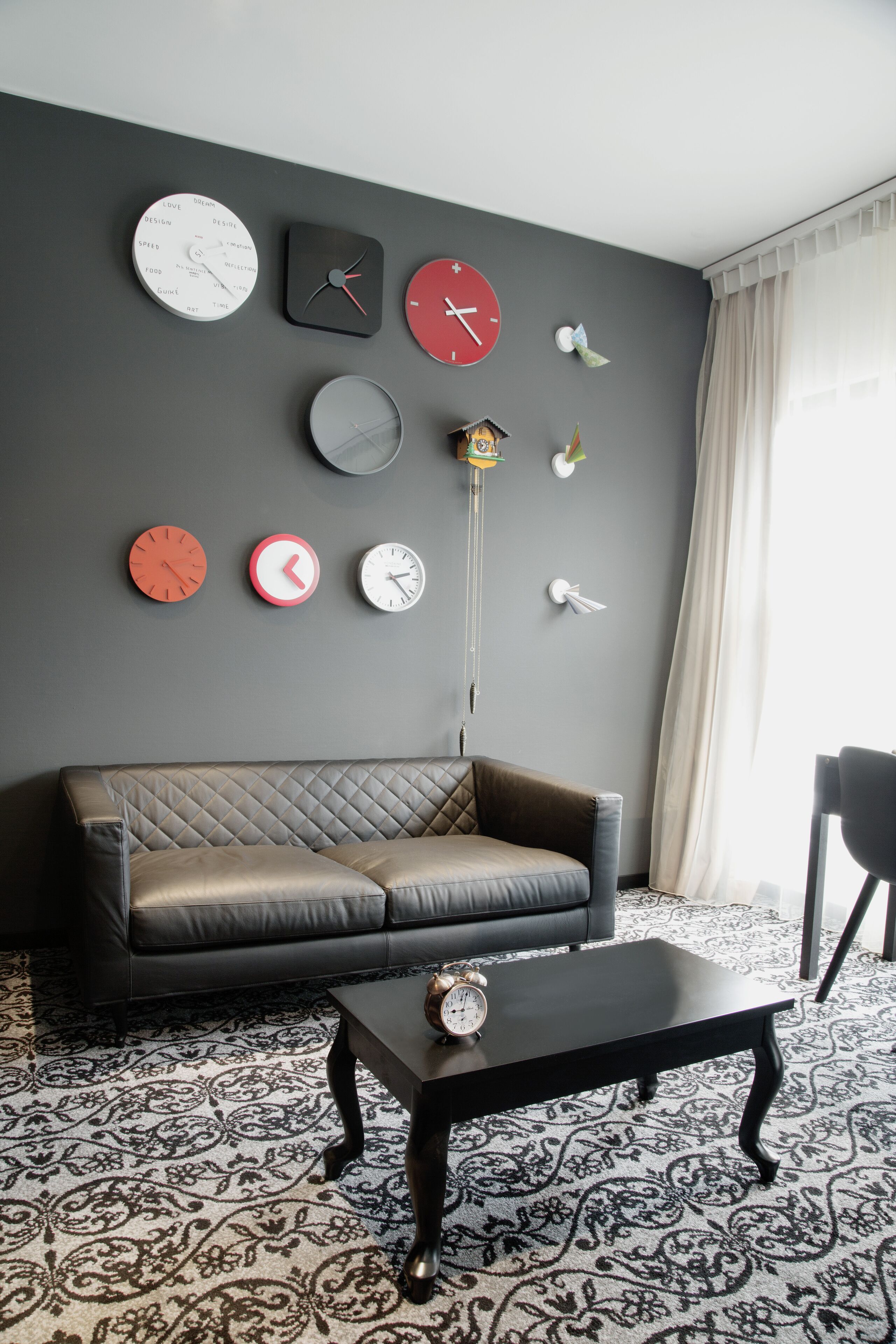 A living room with a black leather sofa, a black coffee table, and a patterned rug. There are multiple clocks on the wall and a hanging plant.