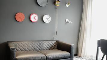 A living room with a black leather sofa, a black coffee table, and a patterned rug. There are multiple clocks on the wall and a hanging plant.