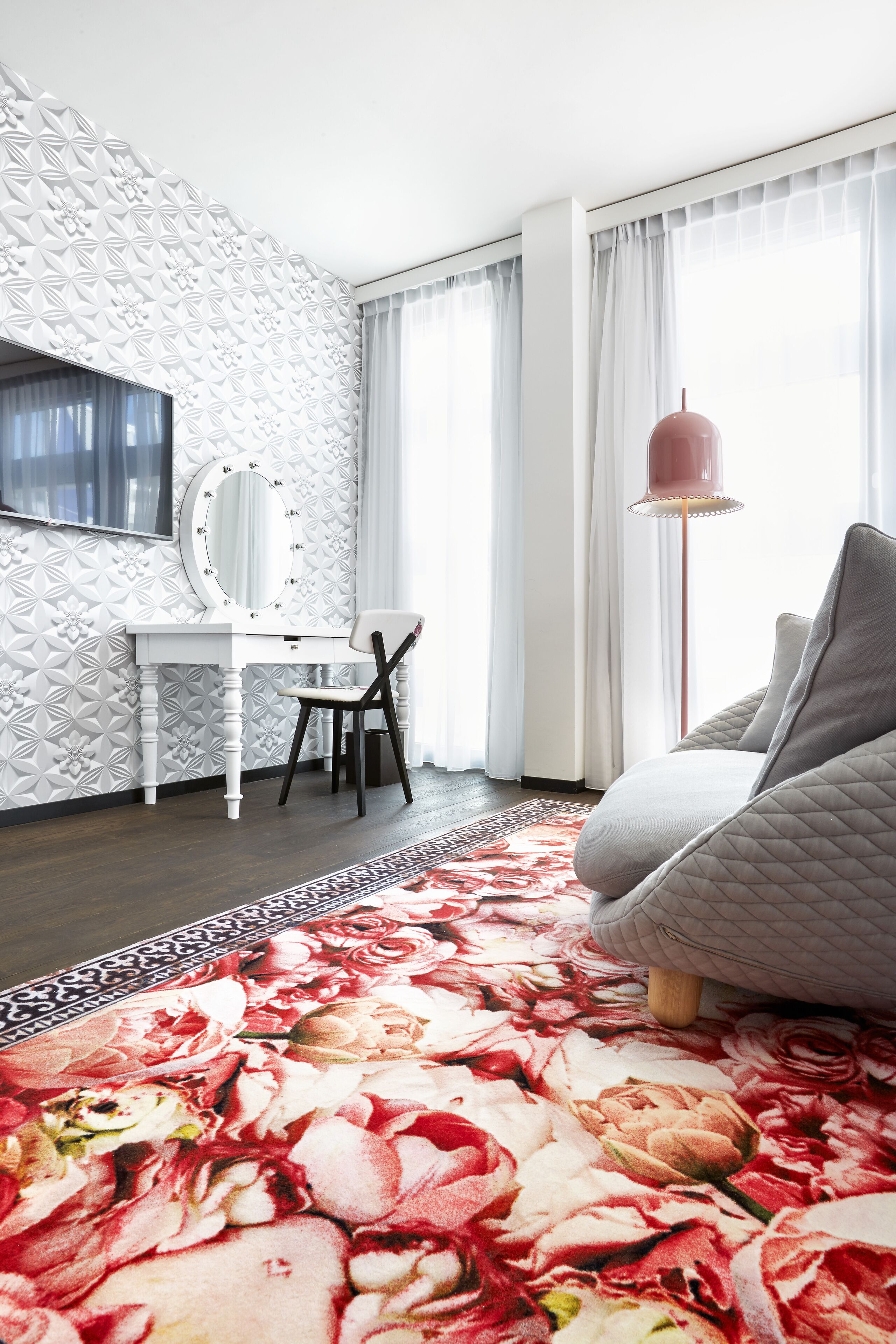 A modern bedroom with a white vanity, a large mirror, a grey sofa, and a floral carpet.