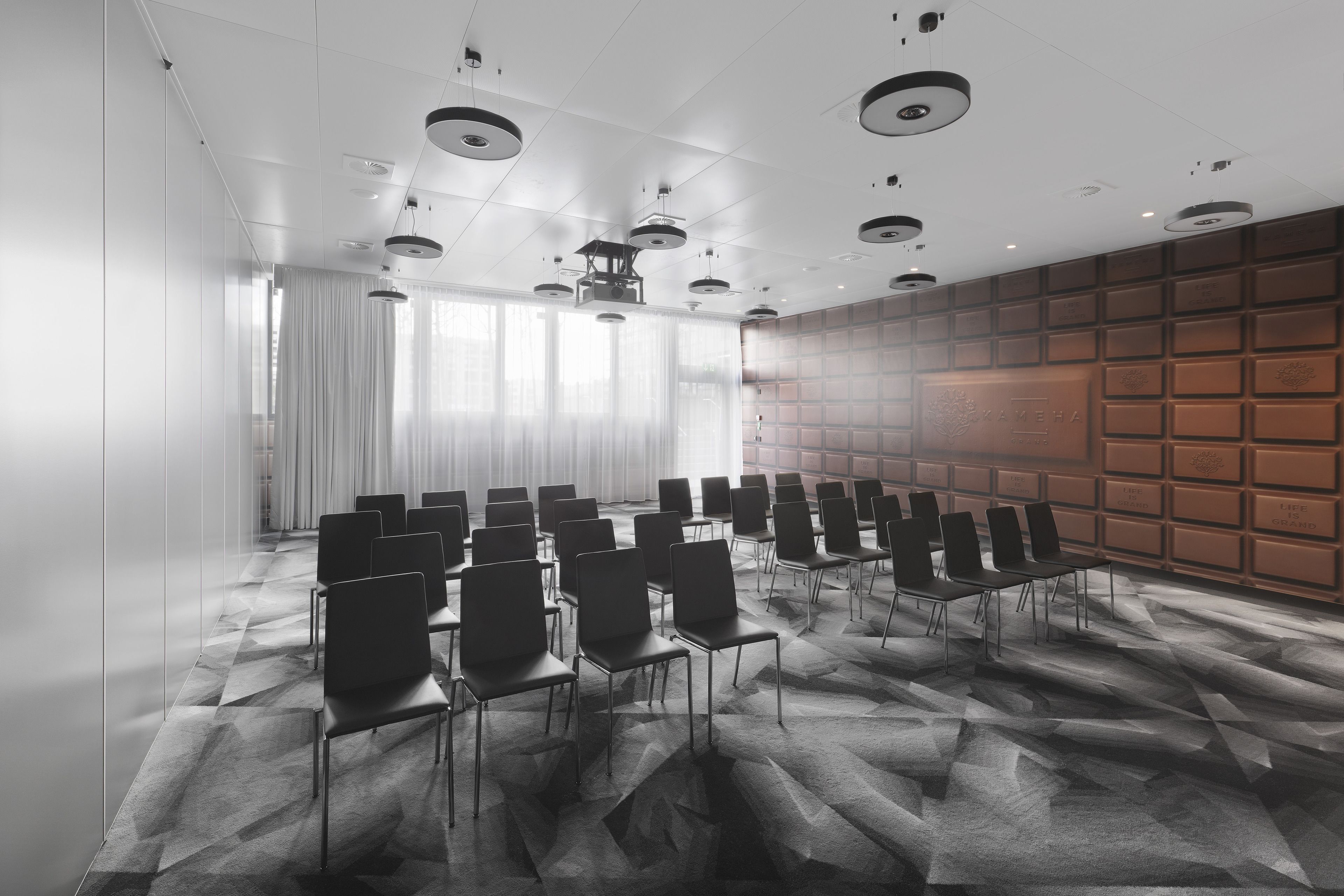 Un salón de conferencias con filas de sillas, un piso con diseño y una pared con paneles organizados en cuadrícula.