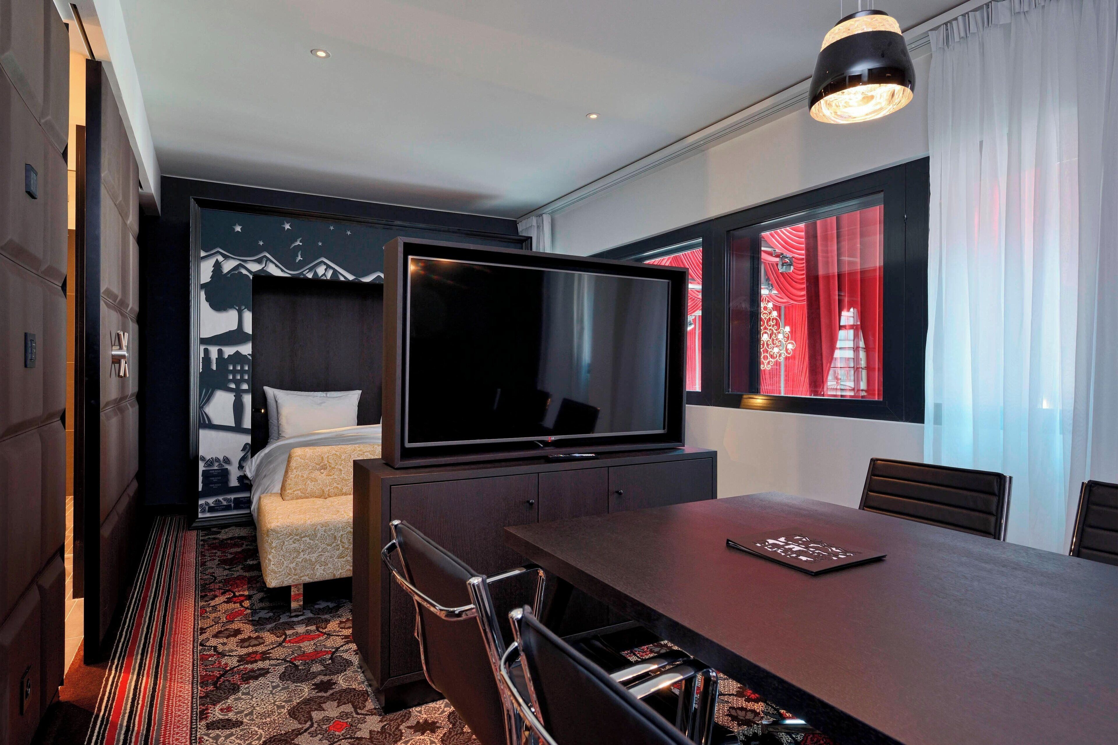 Ein Hotelzimmer mit Bett, Fernseher, Esstisch mit Stühlen und Blick auf die Stadt durch ein Fenster.