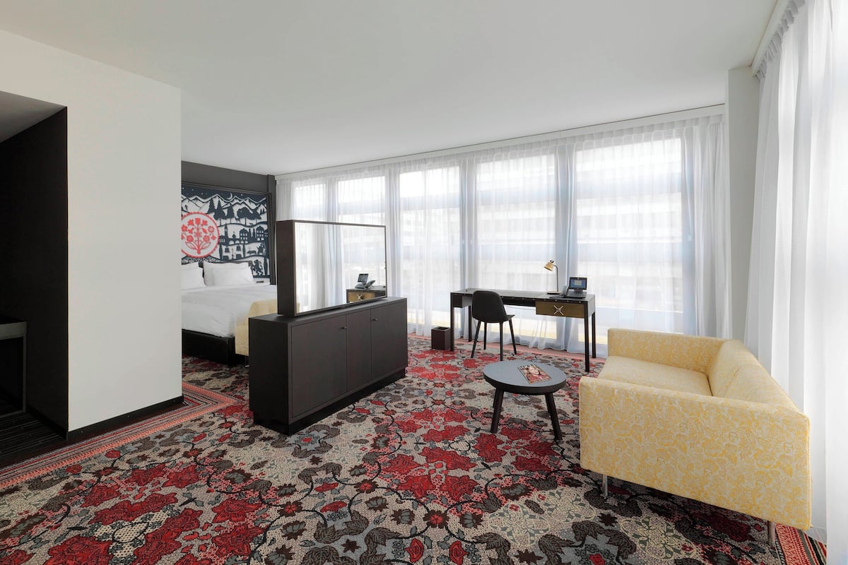 A hotel room with a bed, a desk, a chair, a sofa, and a large window with sheer curtains.