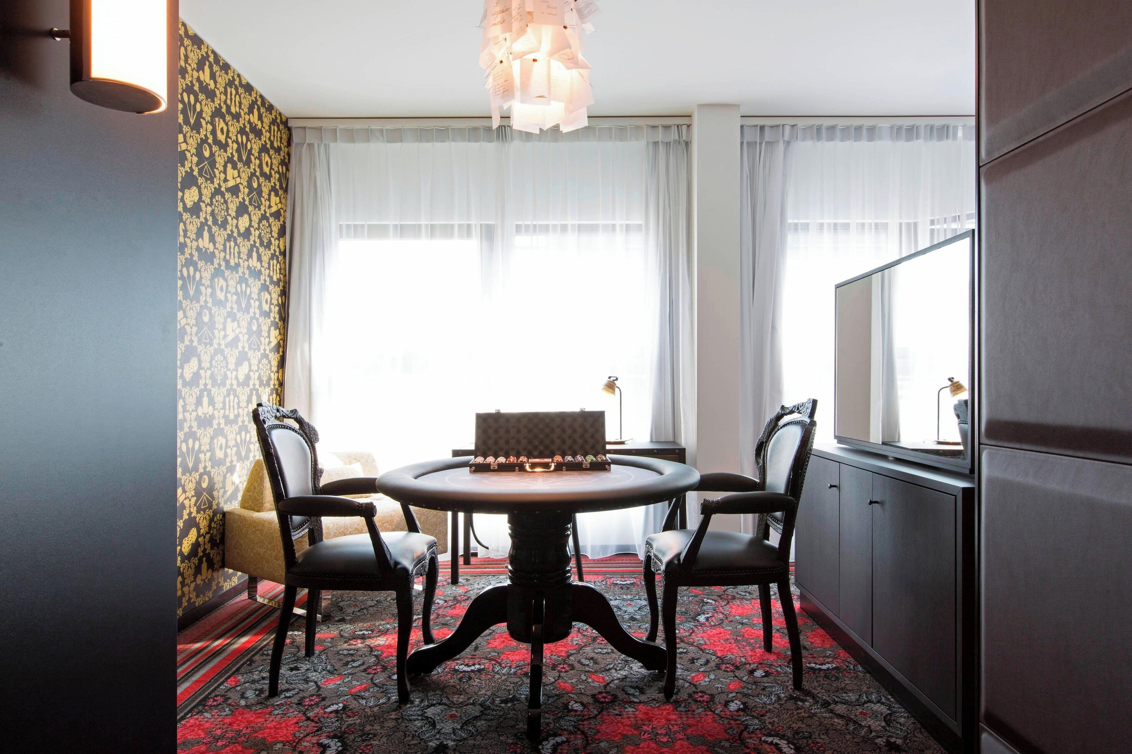 A dining area with a round table, four chairs, a chandelier, and a large window with curtains.
