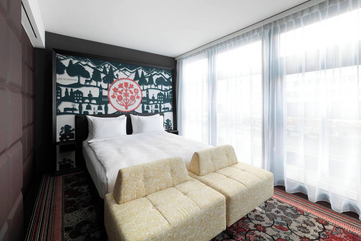 A hotel room with a large bed, two armchairs, a decorative wall art, and a view of the city through sheer curtains.