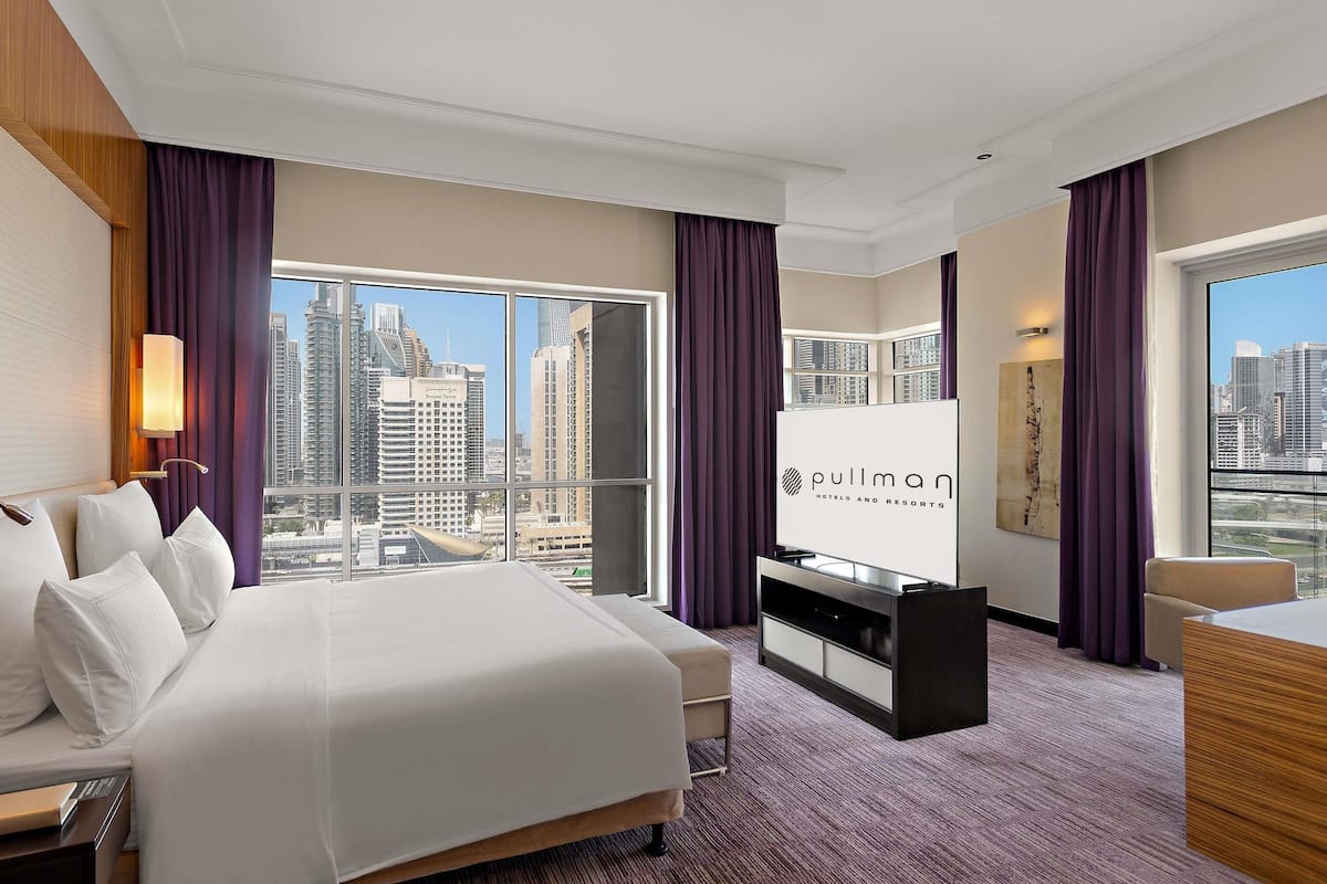 Ein Hotelzimmer mit einem großen Bett, einem Fernseher, einem Schreibtisch und Blick auf die Stadtlandschaft.