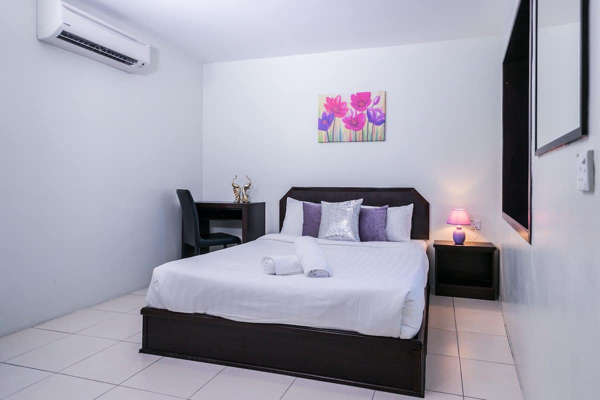 Una cama bien hecha con sábanas blancas, una lámpara de noche y un cuadro de flores en la pared.