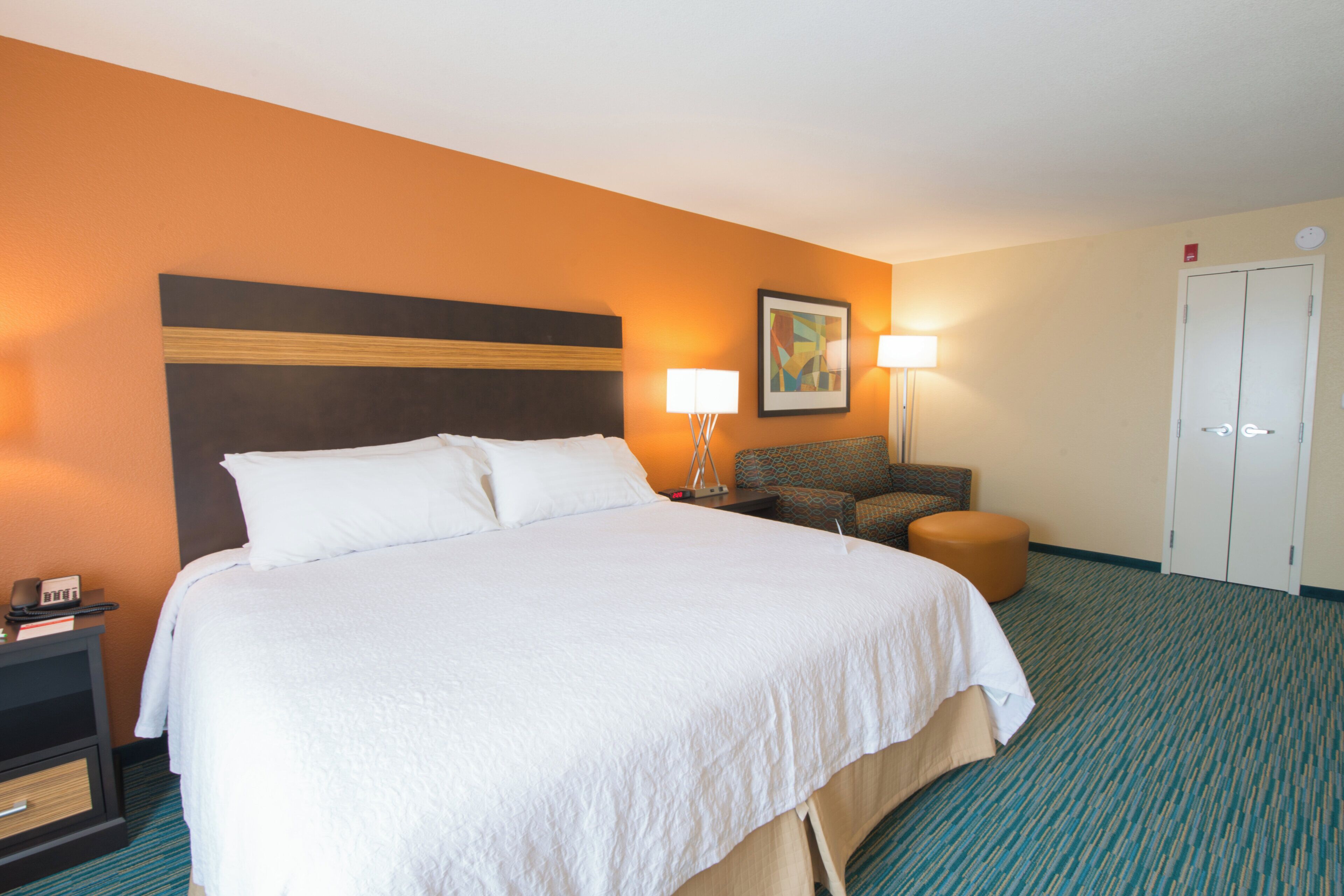 A hotel room with a large bed, a nightstand, a lamp, a chair, and a wardrobe.