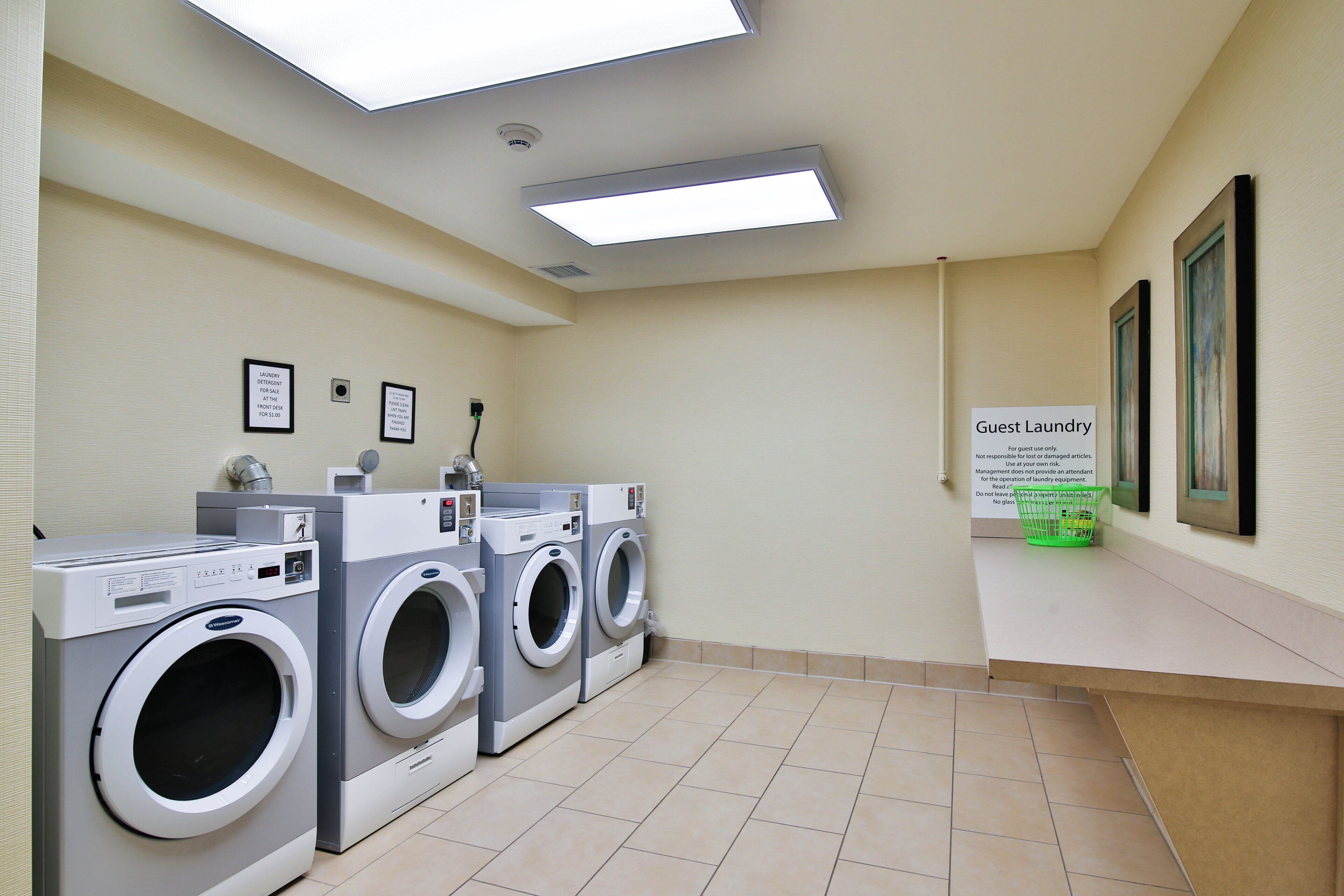 A laundry facility with three front-loading washing machines, a sign indicating guest laundry, and a green basket for laundry items.