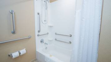 Un baño con bañera, barras de apoyo, un dispensador de papel higiénico y zona de ducha.