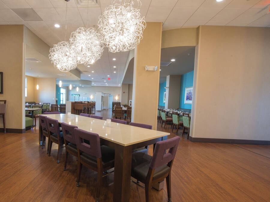 A spacious dining area with a long table and multiple chairs, overhead lighting, and a visible exit sign.