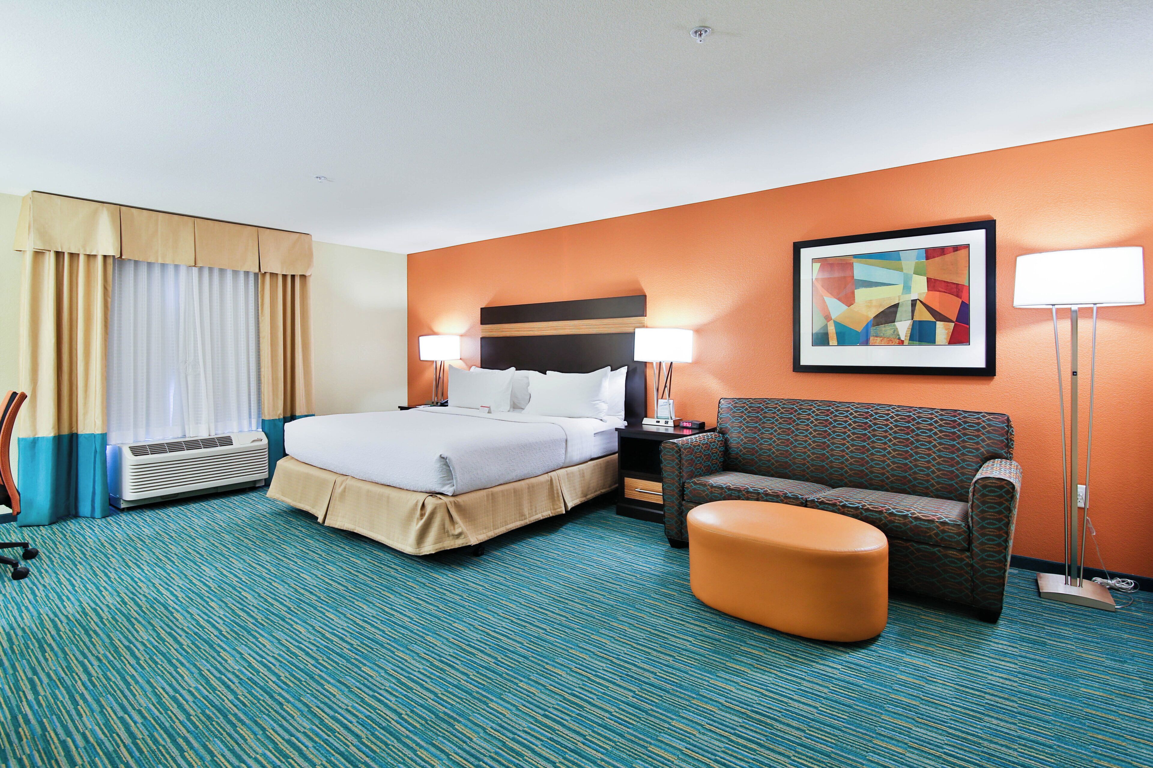 A hotel room with a bed, a chair, a desk, a lamp, and a painting on the wall.