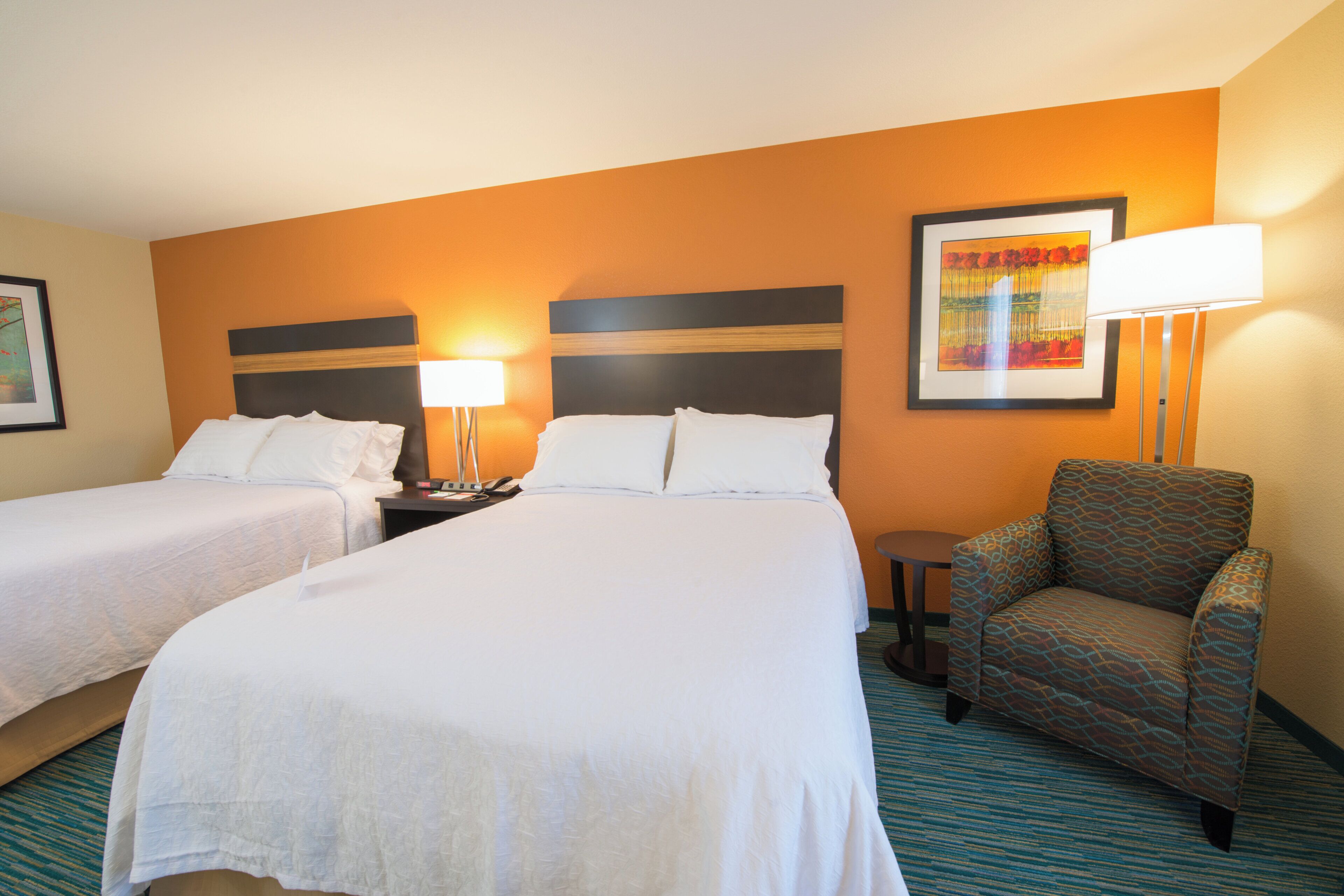 A hotel room with two beds, a chair, a lamp, and a framed picture on the wall.