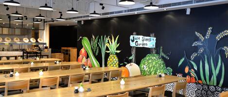 Un restaurant avec des tables et des chaises en bois, une fresque représentant des plantes tropicales et une enseigne sur laquelle on peut lire : « Just Sleep Garden ».
