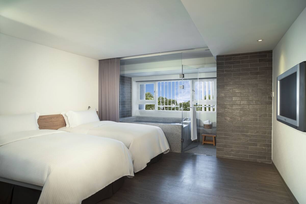 A modern hotel room with two beds, a balcony with a view, and a brick accent wall.