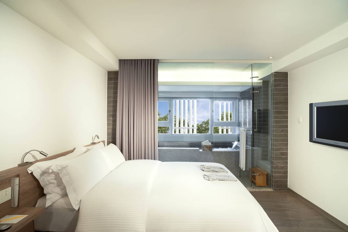 A modern hotel room with a large bed, a TV mounted on the wall, and a glass door leading to a balcony with a view of the sky and trees.