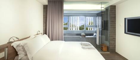 Habitación de hotel moderna con una cama grande, un televisor montado en la pared y una puerta de vidrio que da a un balcón con vistas al cielo y árboles.