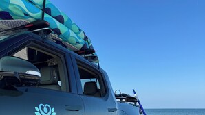 Un SUV fosc estacionat a una platja sorrenca, amb una taula de surf i una taula de rem de color blau sobre la baca.