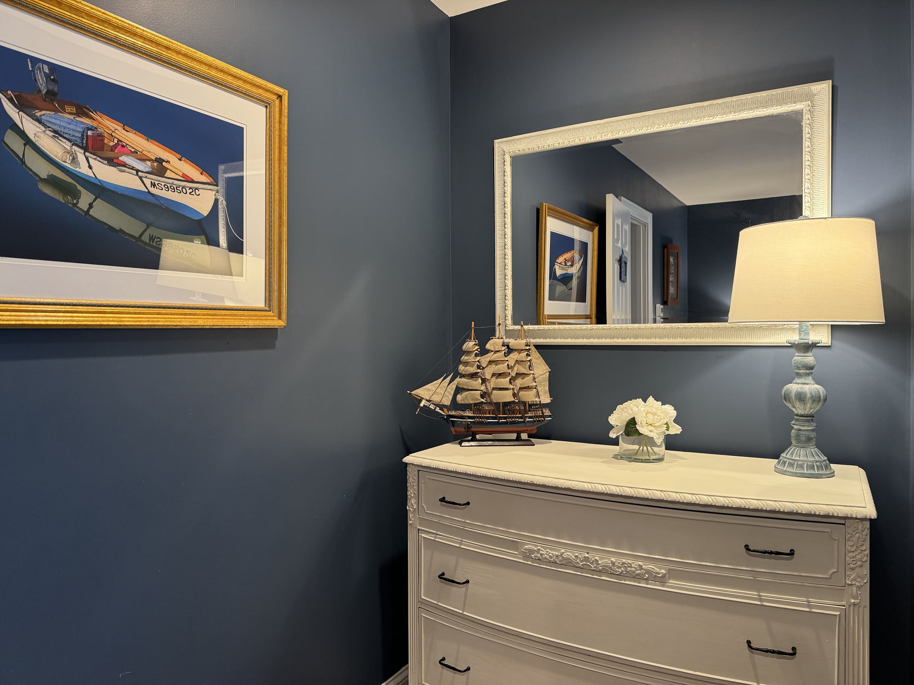 Un bagno con una parete blu navy, un mobile vanitario bianco con tre cassetti, un modellino di nave, un quadro incorniciato raffigurante una barca e uno specchio con cornice dorata.