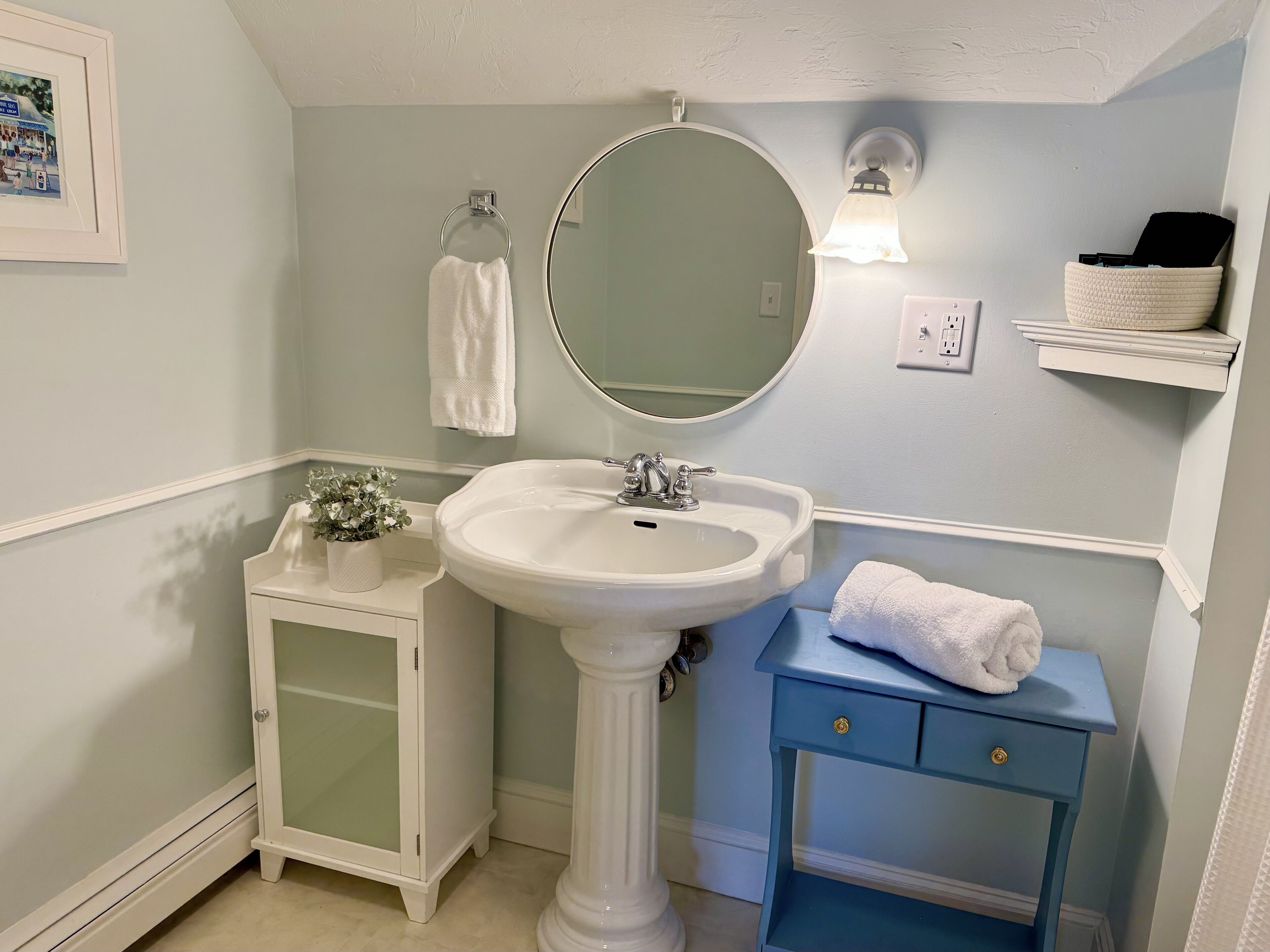 A bathroom with a pedestal sink, a mirror, a small cabinet, a blue vanity, and a shelf with toiletries.