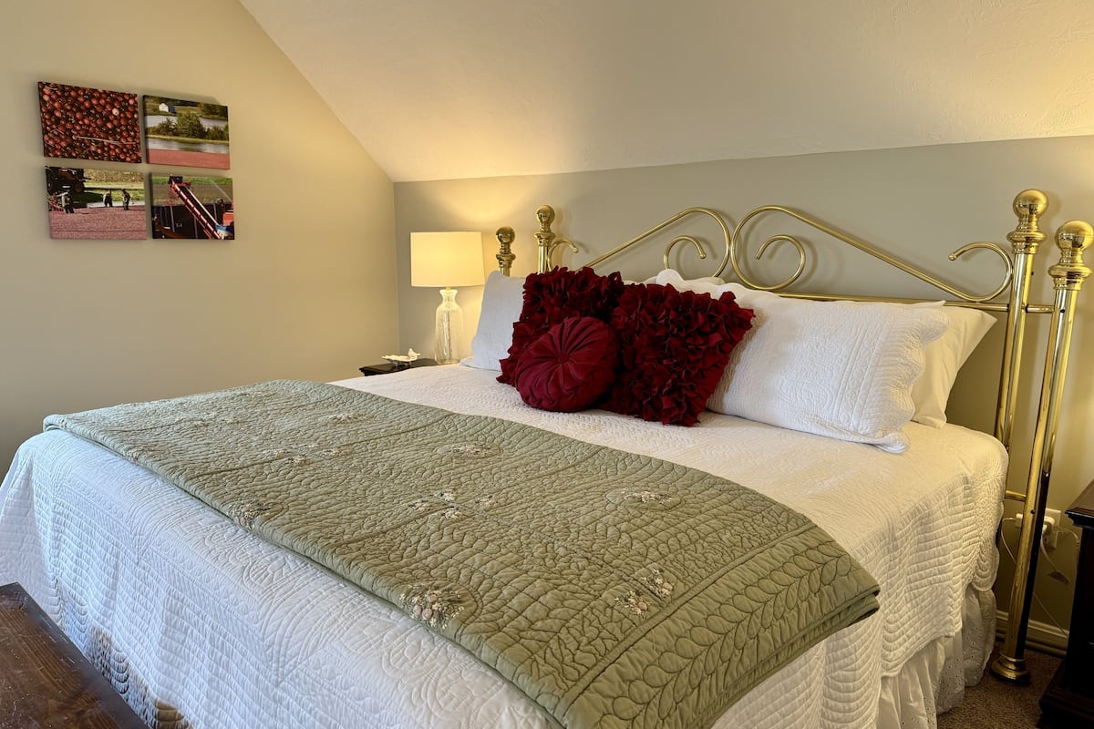 Una cama bien hecha con colcha verde, almohadas blancas y almohadones rojos. Hay una mesita de noche con una lámpara y una pequeña mesa junto a la cama.