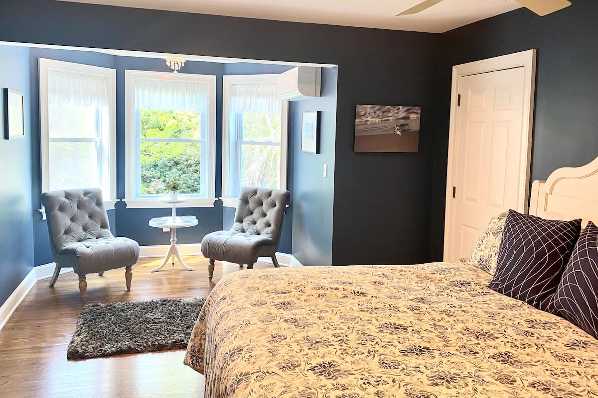 Un dormitorio con una cama, dos sillones, una mesita y una ventana con cortinas.