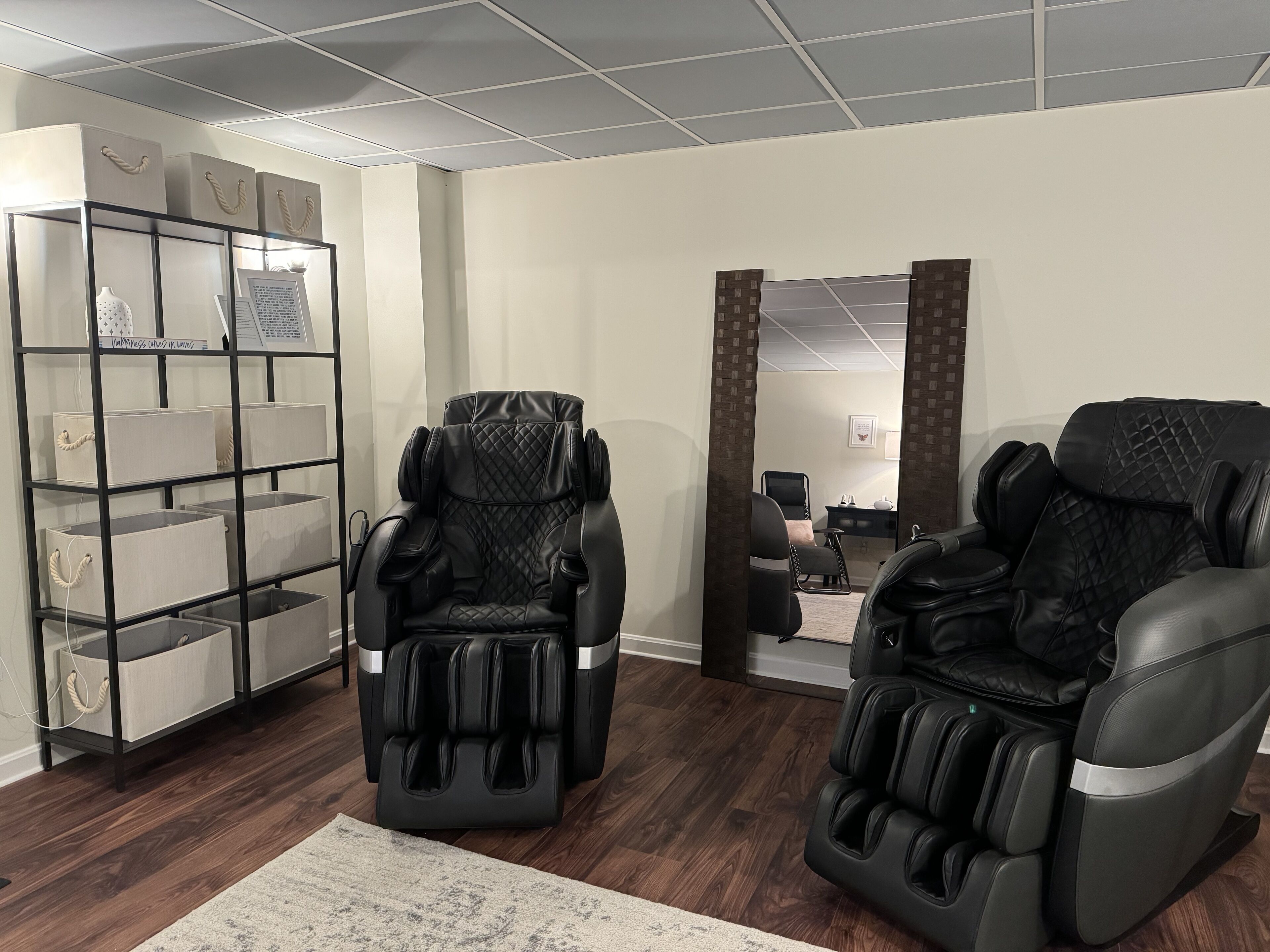 A room with two massage chairs, a storage unit with labeled bins, and a mirror reflecting a desk and chair.