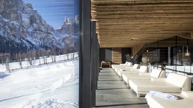 A modern hotel interior with a view of snow-covered mountains through large windows, a row of white sofas, and a wooden ceiling. - Hotel Cappella (Corvara in Badia)