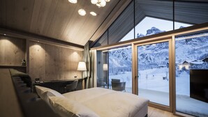 A bedroom with a large bed, wooden ceiling, and a view of a snowy mountain landscape through a glass door. - Hotel Cappella (Corvara in Badia)