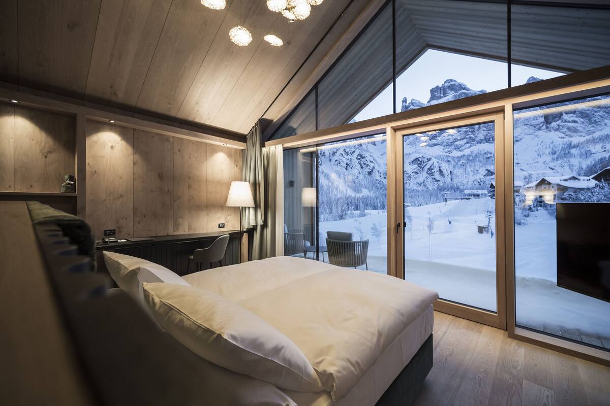 A bedroom with a large bed, wooden ceiling, and a view of a snowy mountain landscape through a glass door.