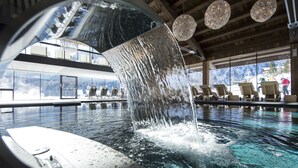 A modern indoor pool with a waterfall feature, lounge chairs, and large windows offering mountain views. - Hotel Cappella (Corvara in Badia)