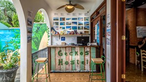 Interior de un hostal con un mostrador de recepción de madera con el letrero "Geckos Hostel", un ventilador de techo, un mural y un piso de baldosas.
