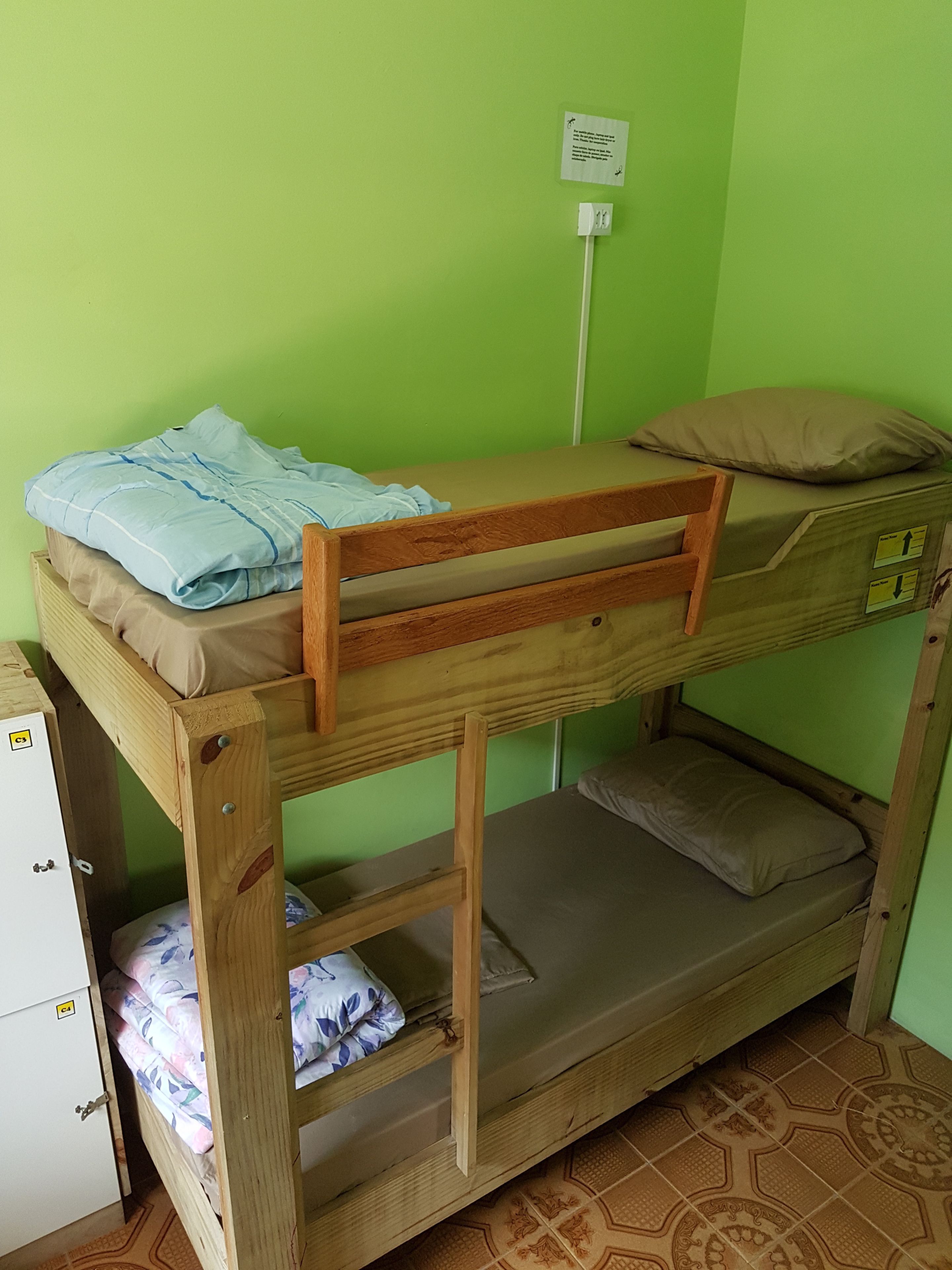 A bunk bed with a wooden frame, a lower bunk with a mattress, and a top bunk with a pillow. There is a white cabinet to the left and a wall socket above the bed.