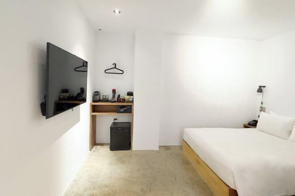 A minimalist hotel room with a bed, a flat-screen TV, a small desk with a chair, and a wall-mounted shelf.
