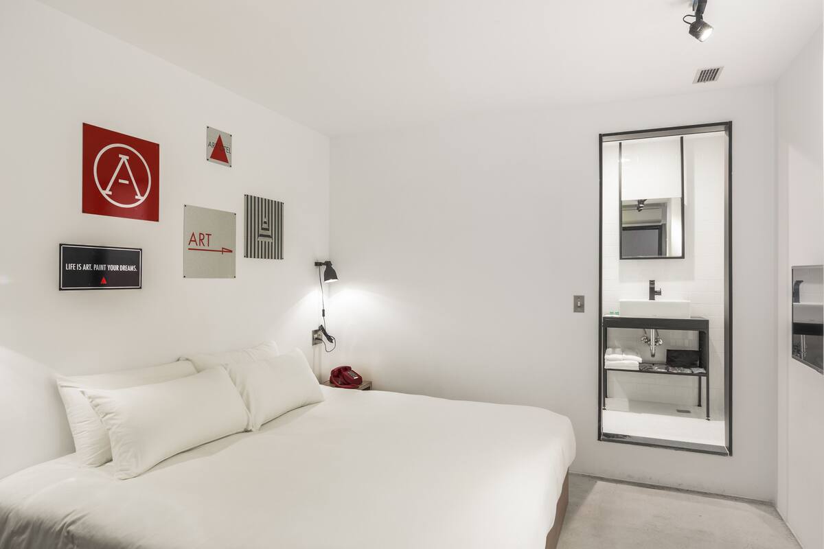 A modern hotel room with a large bed, a bedside table, a red telephone, a wall-mounted mirror, and a bathroom with a sink and mirror.