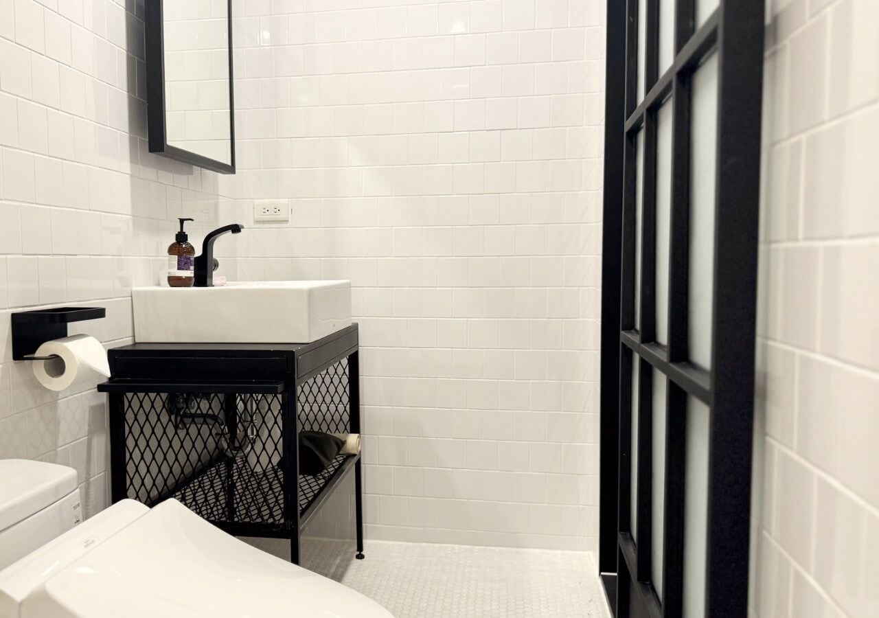 A bathroom with white tiled walls, a black sink with a mirror above it, a toilet, and a shower area with a glass door.
