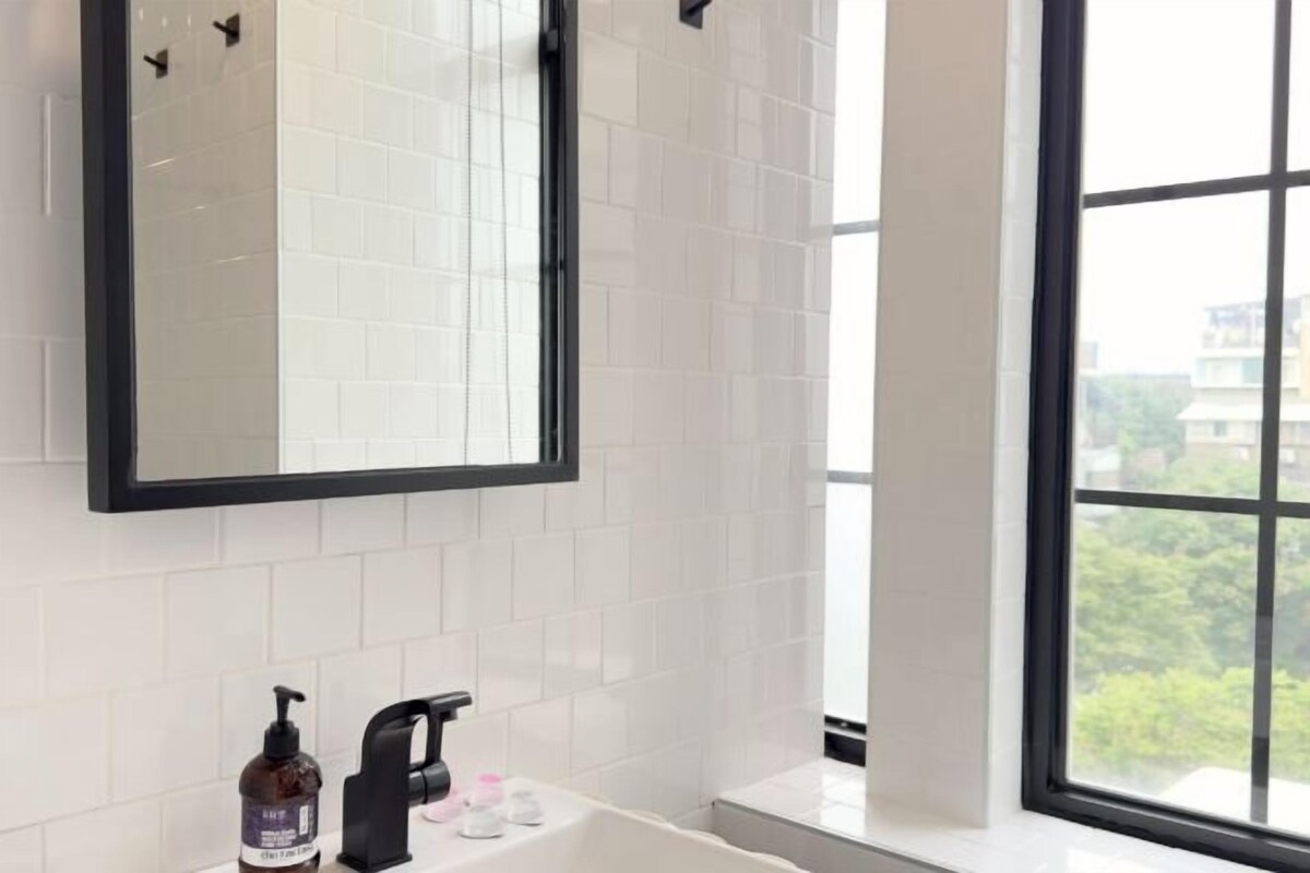 A bathroom with white tiled walls, a black-framed mirror, a white bathtub, and a window with a view of buildings and trees.