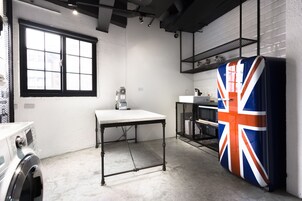 A compact kitchen with a washing machine, a table, a refrigerator with a Union Jack design, and a window with blinds.