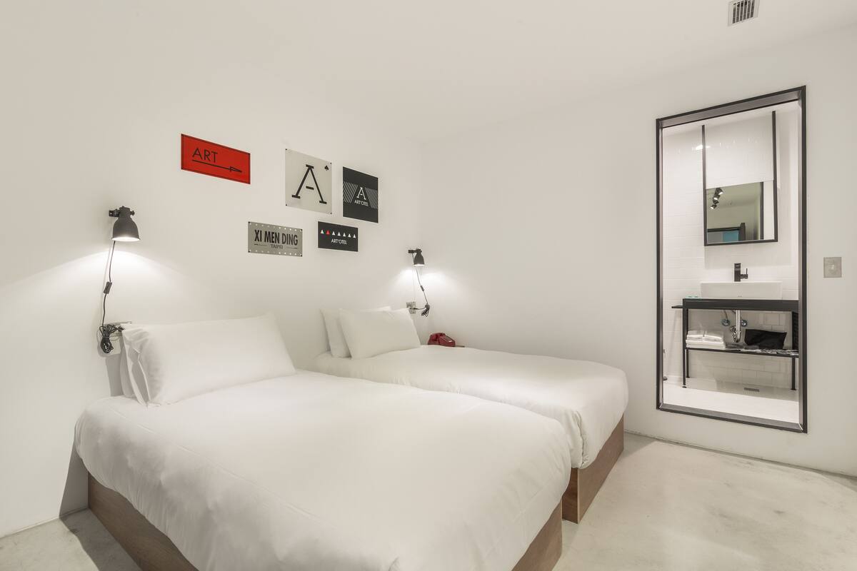 A minimalist bedroom with two beds, a wall-mounted mirror, and a small bathroom with a sink and shelf.