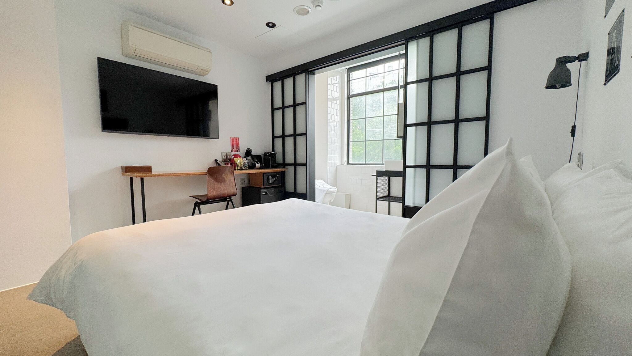 Moderni hotellihuone, jossa on suuri sänky, työpöytä tuolilla, seinälle asennettu taulutelevisio ja suuri ikkuna, josta avautuu näkymä vehreään maisemaan.