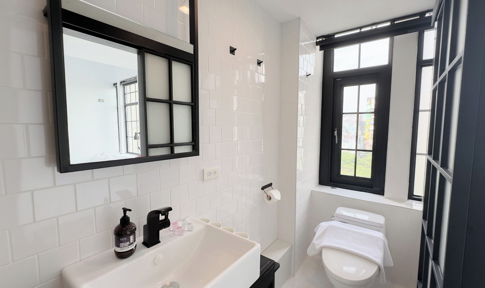 A modern bathroom with white tiles, a large mirror, a bathtub, a toilet, and a window with a view.