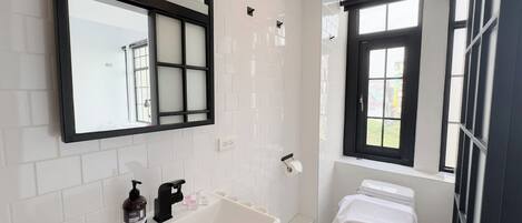 A modern bathroom with white tiles, a large mirror, a bathtub, a toilet, and a window with a view.